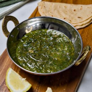 A top-down view of Suva ni Bhaji in a stainless steel bowl with brass handles, served with folded chapatis, fresh garlic cloves, and a lemon wedge on a wooden board.