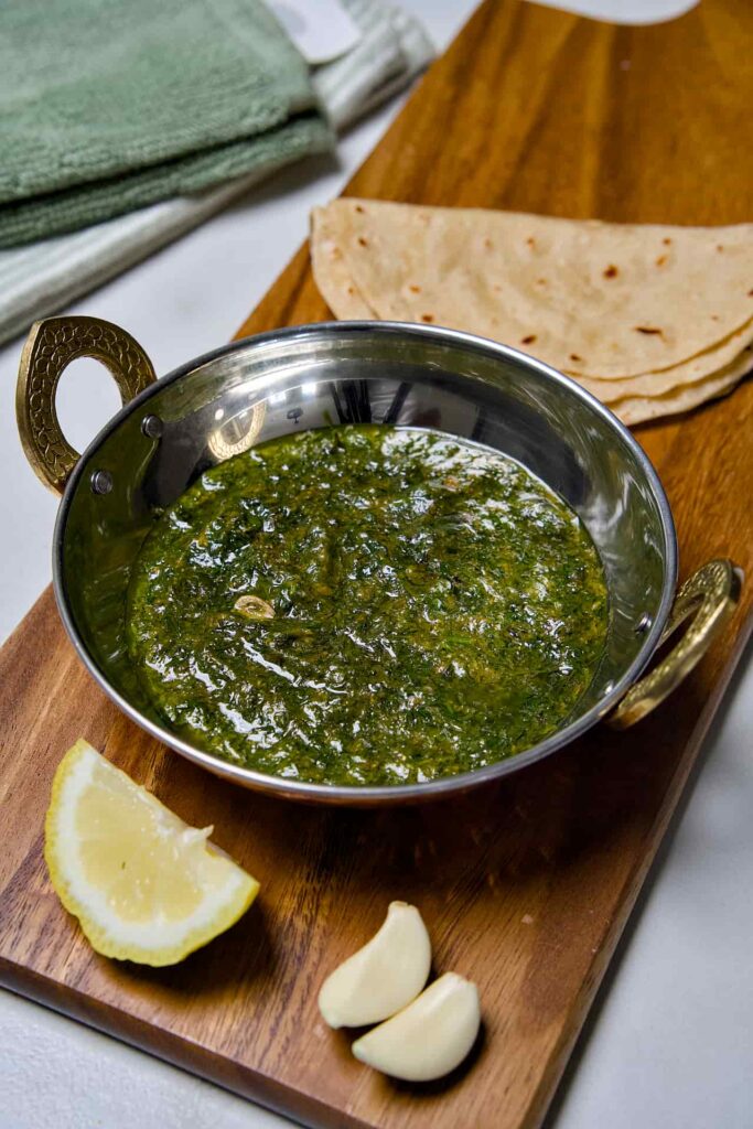 A top-down view of Suva ni Bhaji in a stainless steel bowl with brass handles, served with folded chapatis, fresh garlic cloves, and a lemon wedge on a wooden board.