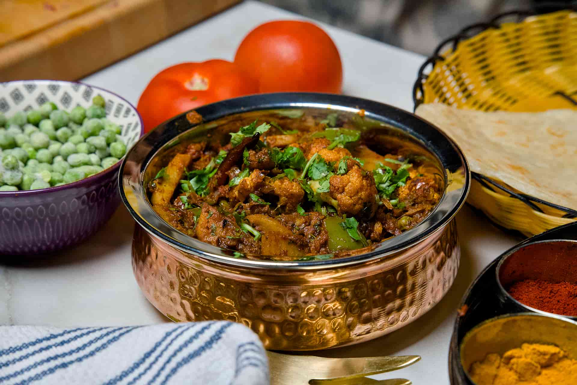 A vibrant bowl of Aloo Gobi Ki Subzi served in a traditional hammered copper handi, garnished with fresh coriander and surrounded by fresh tomatoes and green peas.