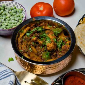 A wide view of the Aloo Gobi curry on a white table, featuring a spice box with turmeric and chili powder, fresh vegetables, and a basket of flatbread in the background.