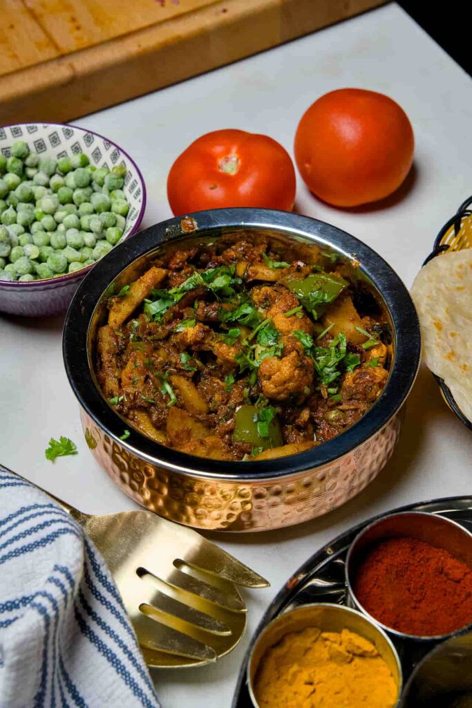 A wide view of the Aloo Gobi curry on a white table, featuring a spice box with turmeric and chili powder, fresh vegetables, and a basket of flatbread in the background.