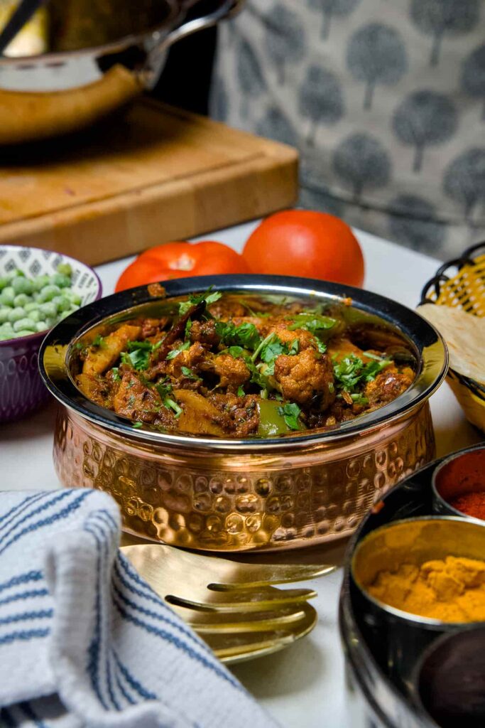 A wide view of the cauliflower potato curry on a table, featuring a spice box with turmeric and chili powder, fresh vegetables, and a basket of roti.