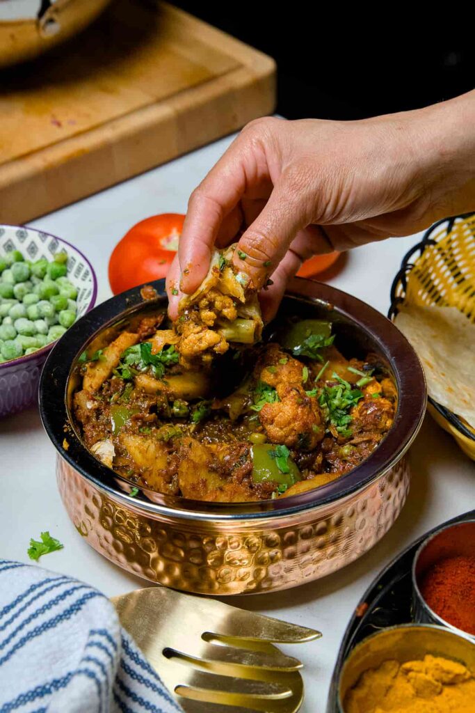 A close-up shot of a hand scooping up a piece of spiced cauliflower and potato from a copper bowl using a piece of warm, folded chapati.