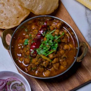 Rich dark chole masala curry served alongside golden bhature bread and seasoned onion salad on a wooden board.