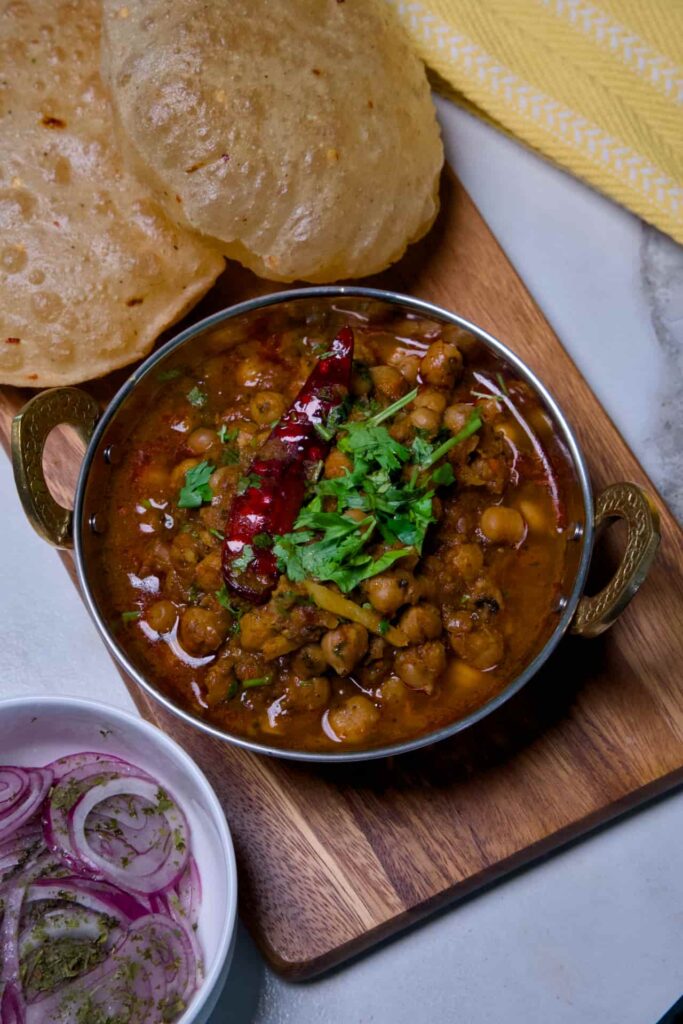 Rich dark chole masala curry served alongside golden bhature bread and seasoned onion salad on a wooden board.