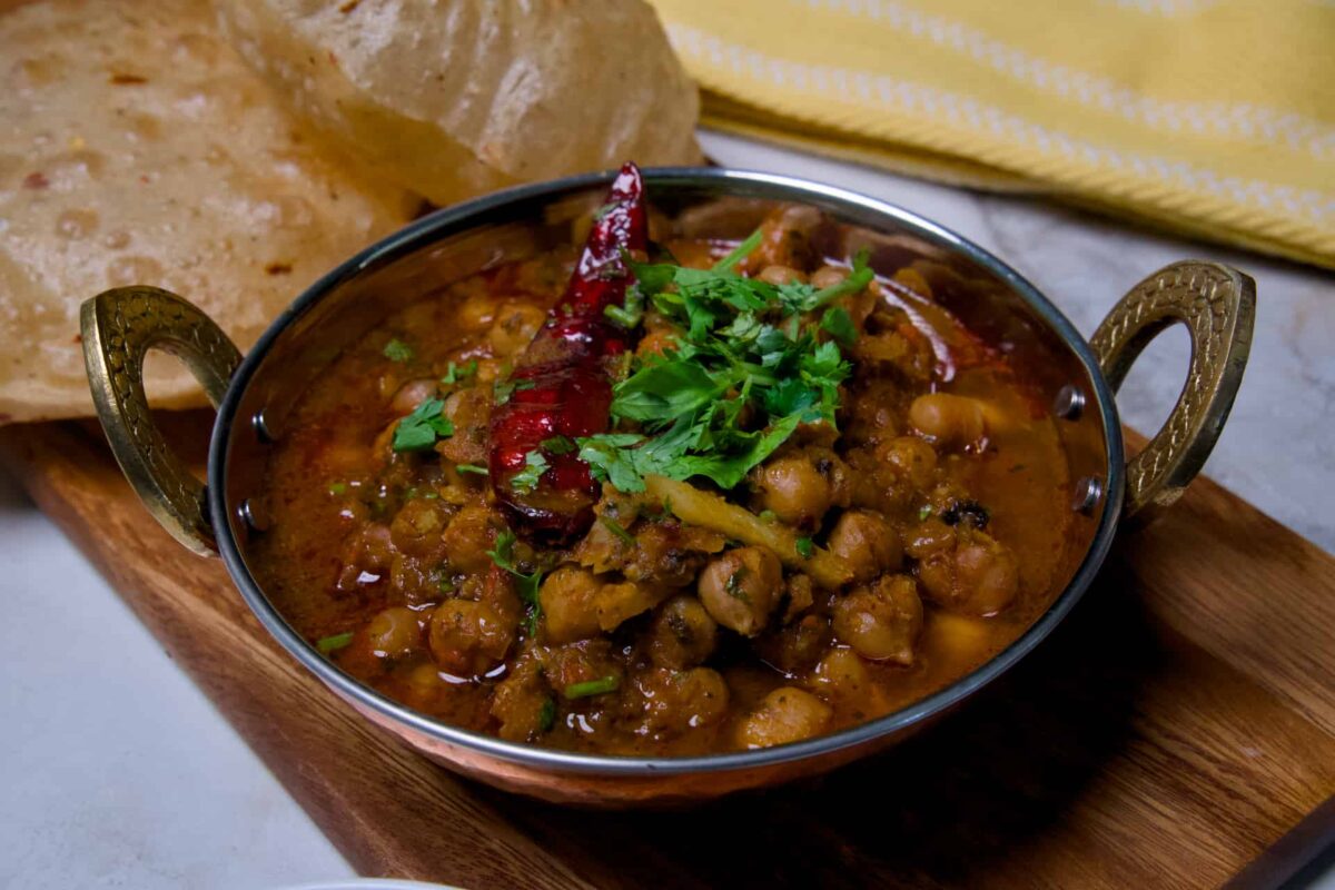 Freshly cooked chole masala in a copper bowl placed on a wooden board with fluffy fried bhature and pickled red onions.