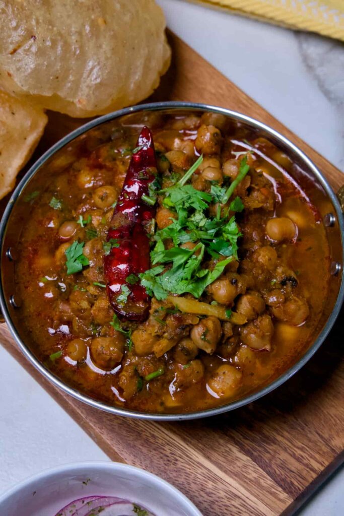 Top-down view of authentic Punjabi Chole Masala in a copper serving bowl topped with cilantro and red chili, served with bhature and sliced onions.